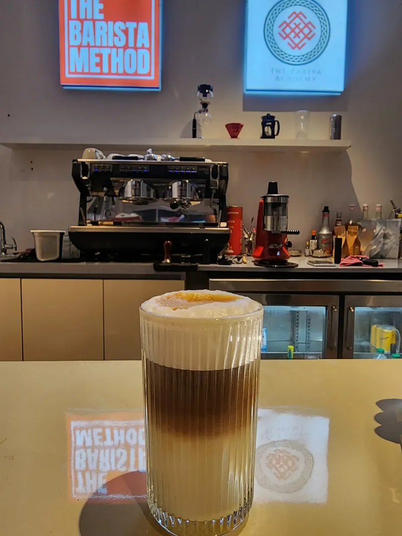 A layered latte in a ribbed glass on the lab counter, with an espresso machine and the Zariya Academy sign behind it